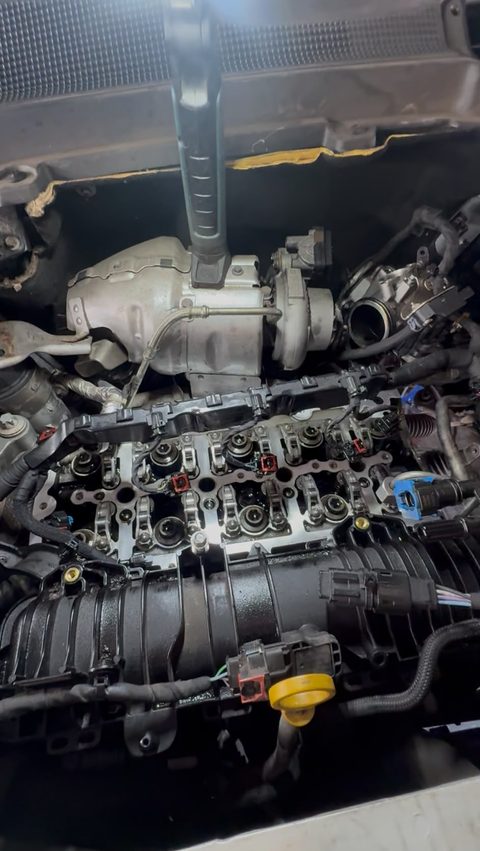 Engine bay with the cam cover off, valve gear and camshaft area exposed, turbo visible and a long tool resting on top, at AGR Performance, Stafford
