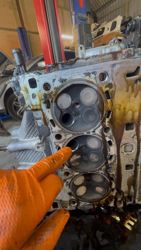 Removed cylinder head, combustion side with valves visible, a gloved finger pointing, workshop ramps behind, at AGR Performance, Stafford