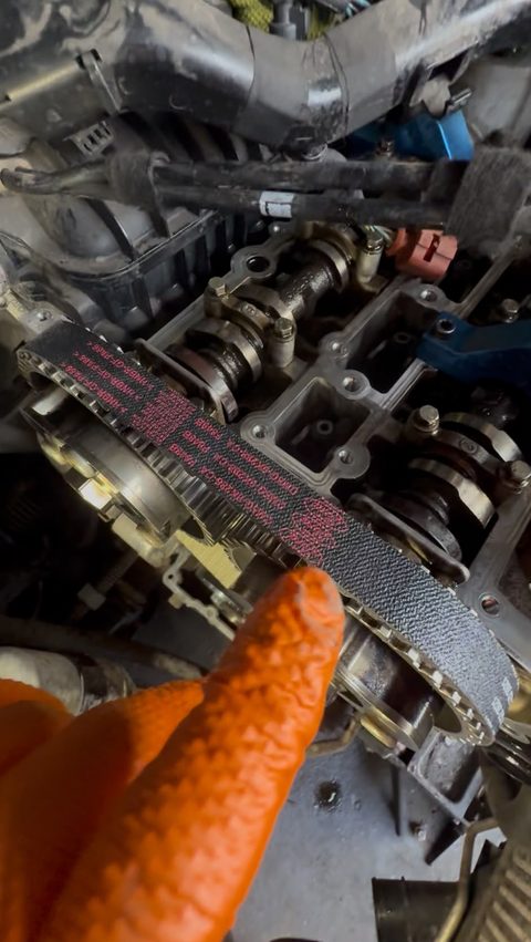 Gloved hand holding the old wet belt on the camshaft pulleys with the cam cover off, at AGR Performance, Stafford