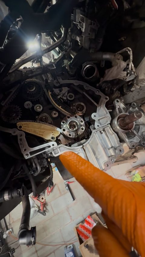 Front of an engine with the timing cover off, sprockets and crank hub exposed, a gloved finger pointing, workshop floor visible, at AGR Performance, Stafford